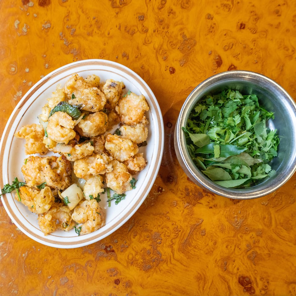 Plate of golden fried vegetable fritters beside a metal bowl of fresh cilantro and leafy herbs on a wooden table.
