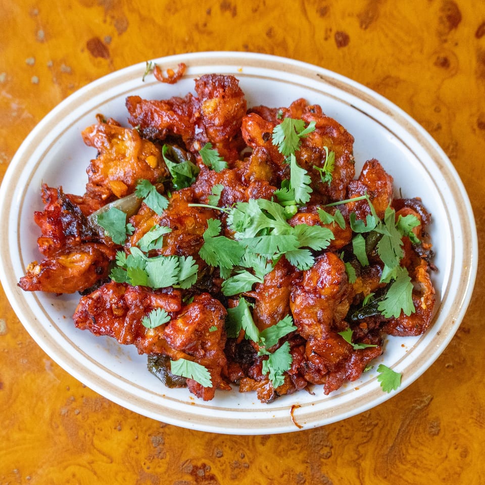 Plate of crispy red-spiced fried chicken garnished with fresh cilantro and curry leaves on a wooden table.