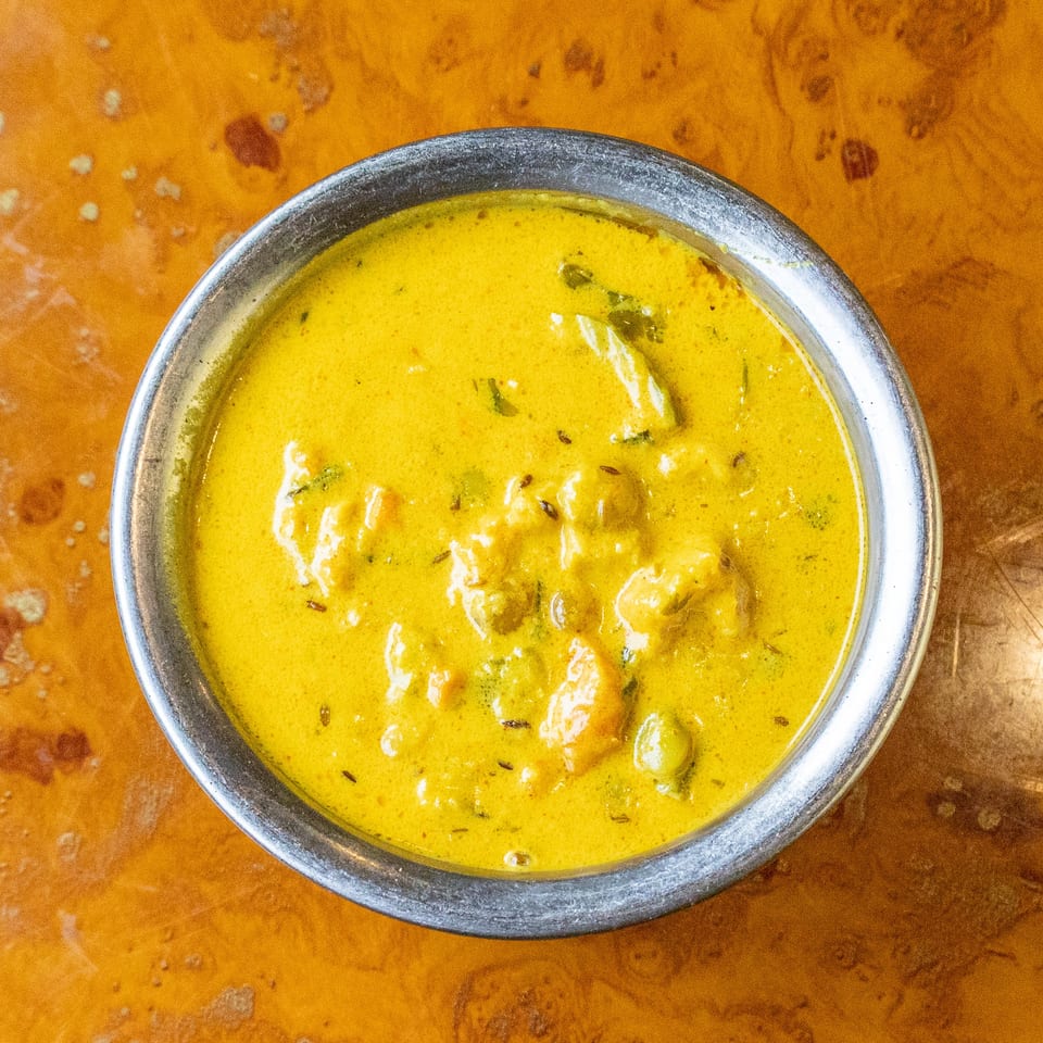 Overhead view of a metal bowl filled with creamy yellow curry garnished with herbs on a rustic wooden table.